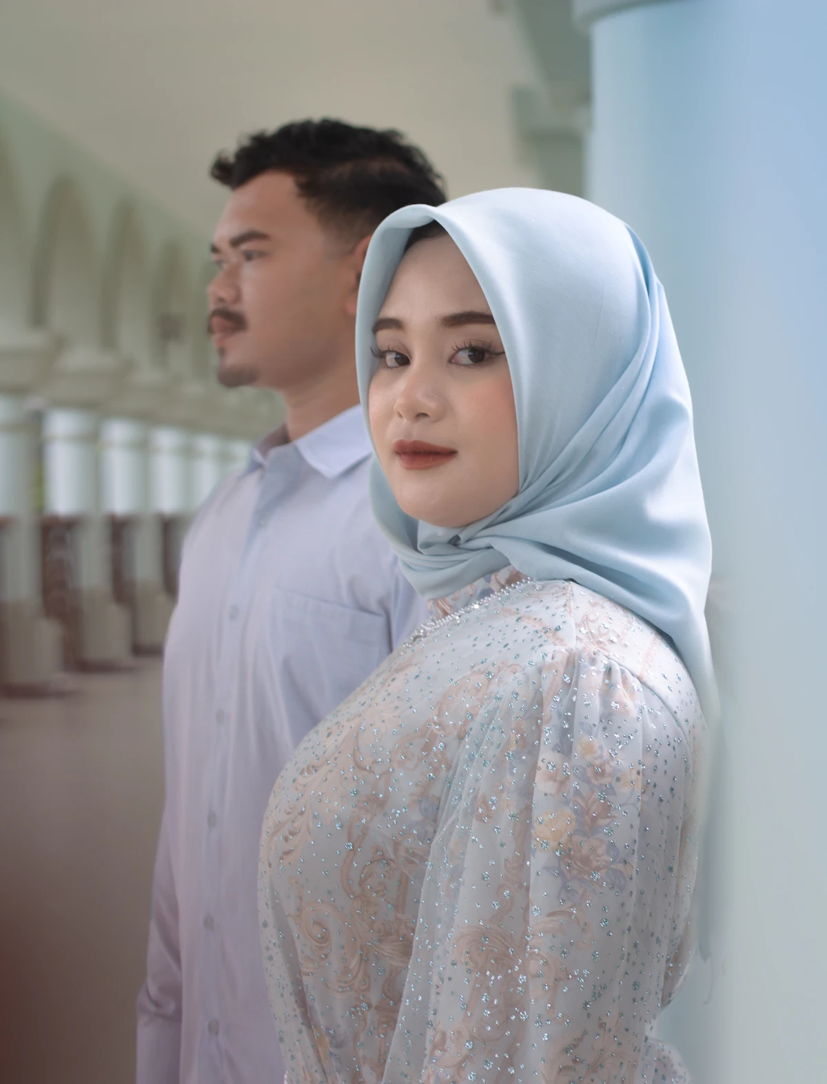 A couple standing in a mosque colonnade, the woman wearing a hijab